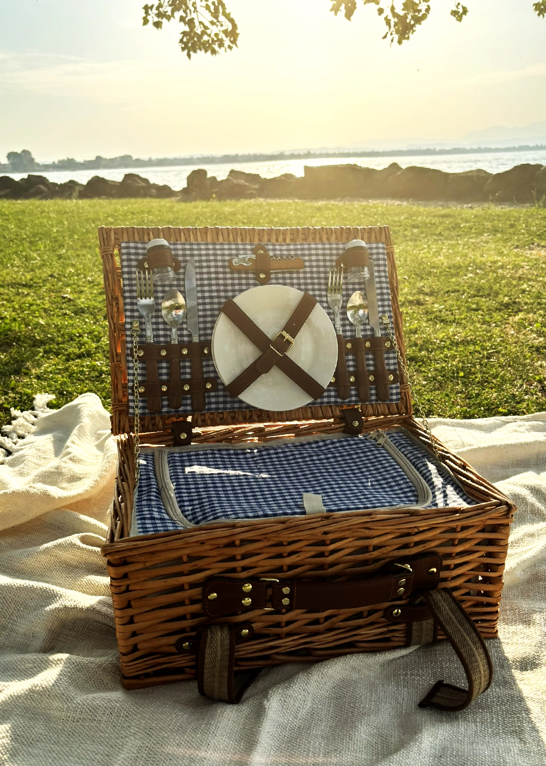 Kit picnic premium aperto con piatti in ceramica, calici e tagliere sul prato, Lago di Garda al tramonto
